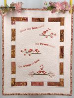 A small light-colored quilt with embroidered birds and phrases in pink tones