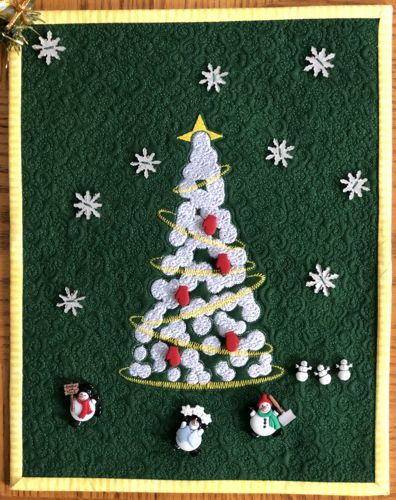 A small forest green quilt with embroidery of a Christmas tree decorated with snowman buttons.