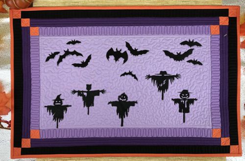 Quilted tablerunner in purple, orange and black fabric with embroidered bats and scarecrows.