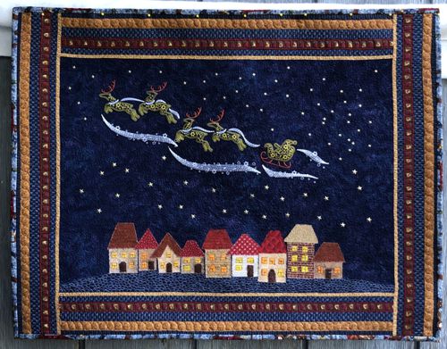 A dark blue wall quilt with embroidered Santa's sleigh in the sky over applique houses