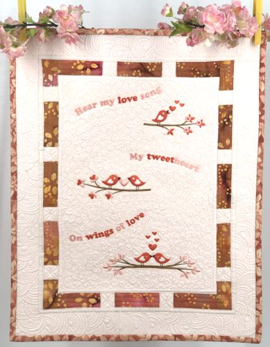 A small light-colored quilt with embroidered birds and phrases in pink tones